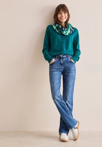 Zelený zapínací kardigan, modré džíny, bílé tenisky s hnědými podrážkami a šál s listovým vzorem. Měkká textura a volný střih.