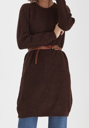 Modèle portant une robe-pull côtelée marron foncé sur un jean bleu, cintrée à la taille par une ceinture beige à clous.