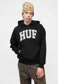 Felpa nera con cappuccio con la scritta "HUF" in caratteri bianchi e grassetto. Tessuto morbido, tasca a marsupio, abbinata a un berretto beige con pois marroni.
