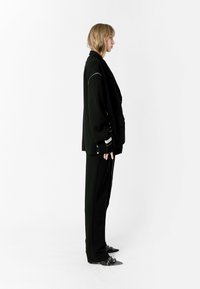 Blazer negro oversize con un diseño estructurado, que presenta líneas de hombros marcadas y un bolsillo frontal. Combinado con pantalones a medida a juego.