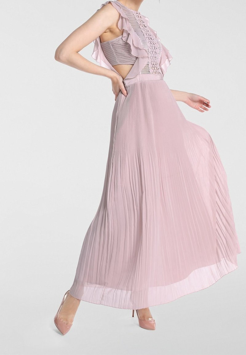 Femme portant une robe maxi rose clair sans manches avec une jupe plissée, un corsage à volants et lacets, et des chaussures à talons hauts couleur chair, posant une main sur la hanche.