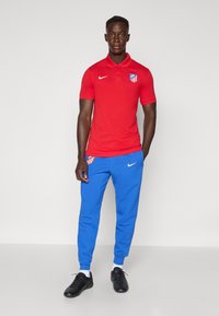 Red polo shirt with collar and Atletico Madrid logo, blue jogger pants with side logo, black sports shoes, and white socks.