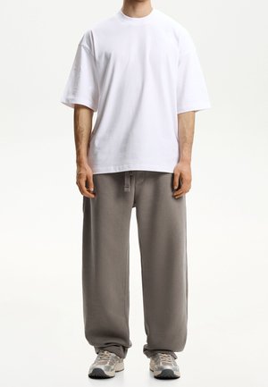 Osoba ubrana w luźny biały t-shirt z krótkim rękawem, szare spodnie dresowe ze sznurkiem i beżowo-szare sneakersy, stojąca na jednolitym tle.