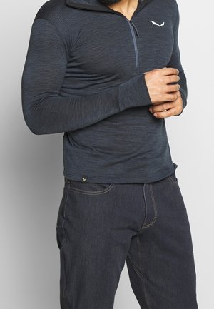 Man wearing a fitted dark blue long-sleeve shirt with a zipper and dark blue jeans, hands clasped near chest.