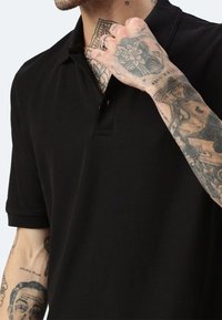 Polo nero in tessuto strutturato, con colletto a due bottoni e maniche corte, indossato da una persona con tatuaggi visibili sull'avambraccio.