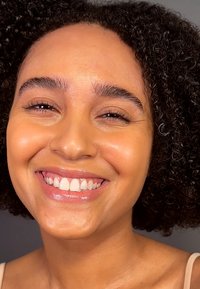 Portrett av en person med krøllete svart hår, glatt hud og et bredt smil som viser hvite tenner. Varm hudtone og naturlig sminke.