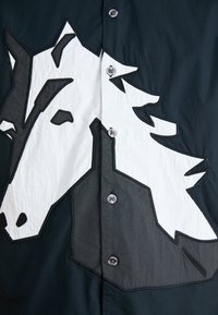 Black shirt featuring a large black and white horse head appliqué. Smooth texture with plastic buttons and contrasting stitching.