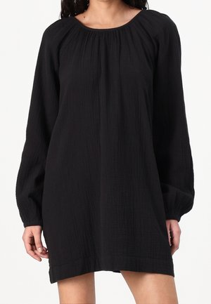 Femme portant une robe mini noire ample à manches longues avec encolure froncée et poignets élastiques sur fond blanc.
