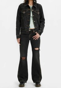 Chaqueta de mezclilla negra con bolsillos de solapa, combinada con jeans acampanados negros desgastados que presentan agujeros pronunciados en las rodillas. Llevada con una blusa ligera de estampado.