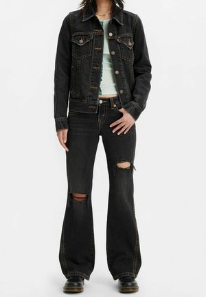Giacca di jeans nera con tasche a toppa, abbinata a jeans neri svasati e usurati con evidenti strappi sulle ginocchia. Indossata con un top chiaro a fantasia.