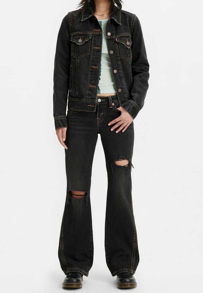 Chaqueta de mezclilla negra con bolsillos de solapa, combinada con jeans acampanados negros desgastados que presentan agujeros pronunciados en las rodillas. Llevada con una blusa ligera de estampado.