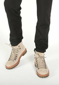 Botas de senderismo de ante beige con suelas gruesas marrones, llevadas con pantalones negros remangados sobre un fondo blanco.