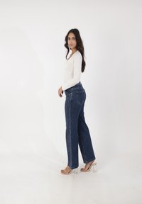 Vit långärmad topp i kombination med högmidjade, blå denim flare jeans som har sidostygn och genomskinliga klacksandaler.