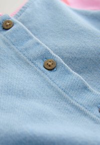 Lyseblåt denimstof med en glat struktur, der har brune knapper syet på forsiden og en pink accentkontur langs kanten.