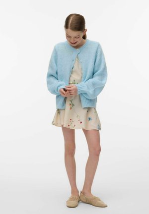 Jeune fille regardant vers le bas, portant un gilet en tricot bleu clair sur une robe fleurie crème avec des chaussures beige tissées sans lacets.