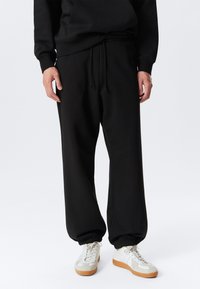 Pantalon de survêtement noir à coupe décontractée, avec des poignets élastiques et une taille à cordon de serrage. Associé à des baskets blanches et beige avec des lacets.