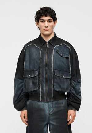 Άνδρας που φοράει σκούρο τζιν μπουφάν με φθαρμένες άκρες, μεγάλες τσέπες με καπάκια και αντίστοιχο τζιν παντελόνι σε απλό φωτεινό φόντο.