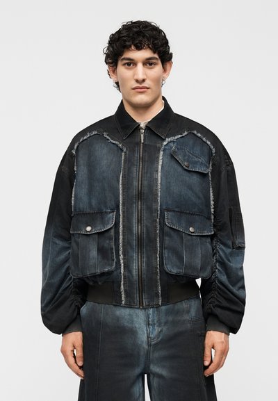 Homme portant une veste en denim foncé avec bords effilochés, grandes poches à rabat et pantalon en denim assorti sur un fond clair uni.