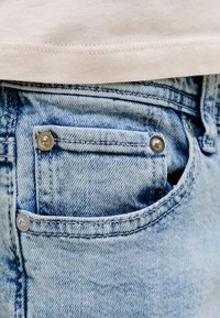 Κοντινή λήψη τσέπης από ανοιχτό μπλε τζιν με μεταλλικά καρφιά, εν μέρει καλυμμένη από λευκό ύφασμα ραφής.