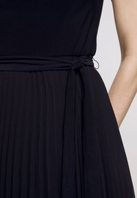 Černé plisované šaty s látkovým páskem uvázaným do mašle v pase, odhalující stranu odkryté paže.