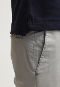 Widok z boku osoby w beżowych spodniach z przednią kieszenią oraz granatowej, teksturowanej koszuli z bocznym rozcięciem.