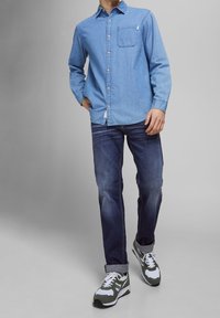 Camisa de mezclilla de botón de color azul claro con un bolsillo en el pecho, combinada con jeans de color azul oscuro y zapatillas gris-verdosas con acentos blancos.