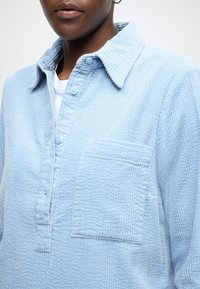 Cămașă de catifea albastru deschis cu guler cu nasturi, buzunar simplu la piept și material texturat; prezintă un model nervurat vizibil.