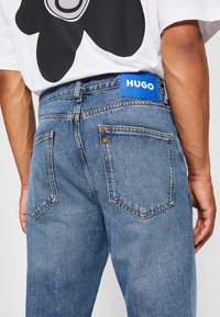 Light blue denim jeans featuring a visible blue HUGO label on the waistband, orange stitching, and classic five-pocket design.