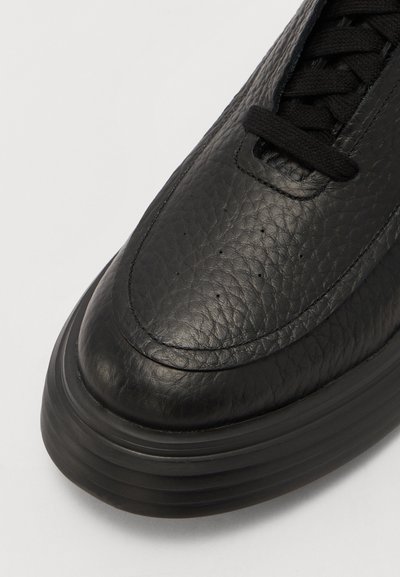 Adidași negri din piele, cu material texturat, vârf rotund și șireți subțiri. Dispune de o talpă din cauciuc negru cu dungi orizontale.