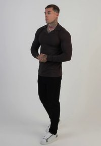 Donkerbruin poloshirt met lange mouwen, zwarte broek en witte sneakers. Heeft een kraag en een nauwsluitend ontwerp, dat de lichaamsvorm accentueert.