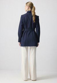 Námornícka modrá košeľa s dlhým rukávom a opaskom v páse, s golierom a manžetami na gombíky, v kombinácii s krémovými voľnými nohavicami.