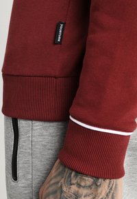 Sudadera burdeos con puños acanalados y un acento blanco, combinada con joggers grises que tienen un bolsillo lateral y un zipper. La etiqueta dice "YOURTURN."