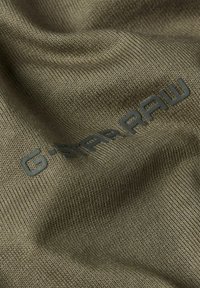 Il tessuto verde oliva di G-Star RAW con una texture liscia presenta un logo in rilievo di una tonalità più scura, parzialmente visibile rispetto al materiale.