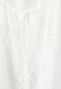 Tissu blanc avec broderie œillet, une patte centrale avec des boutons, et une fermeture à cordon noué près de l'encolure.