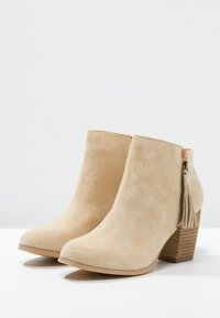 Beige suède enkellaarsjes met een spitse neus, een rits aan de zijkant en een kwastje; vierkante houten hak voor extra stabiliteit.