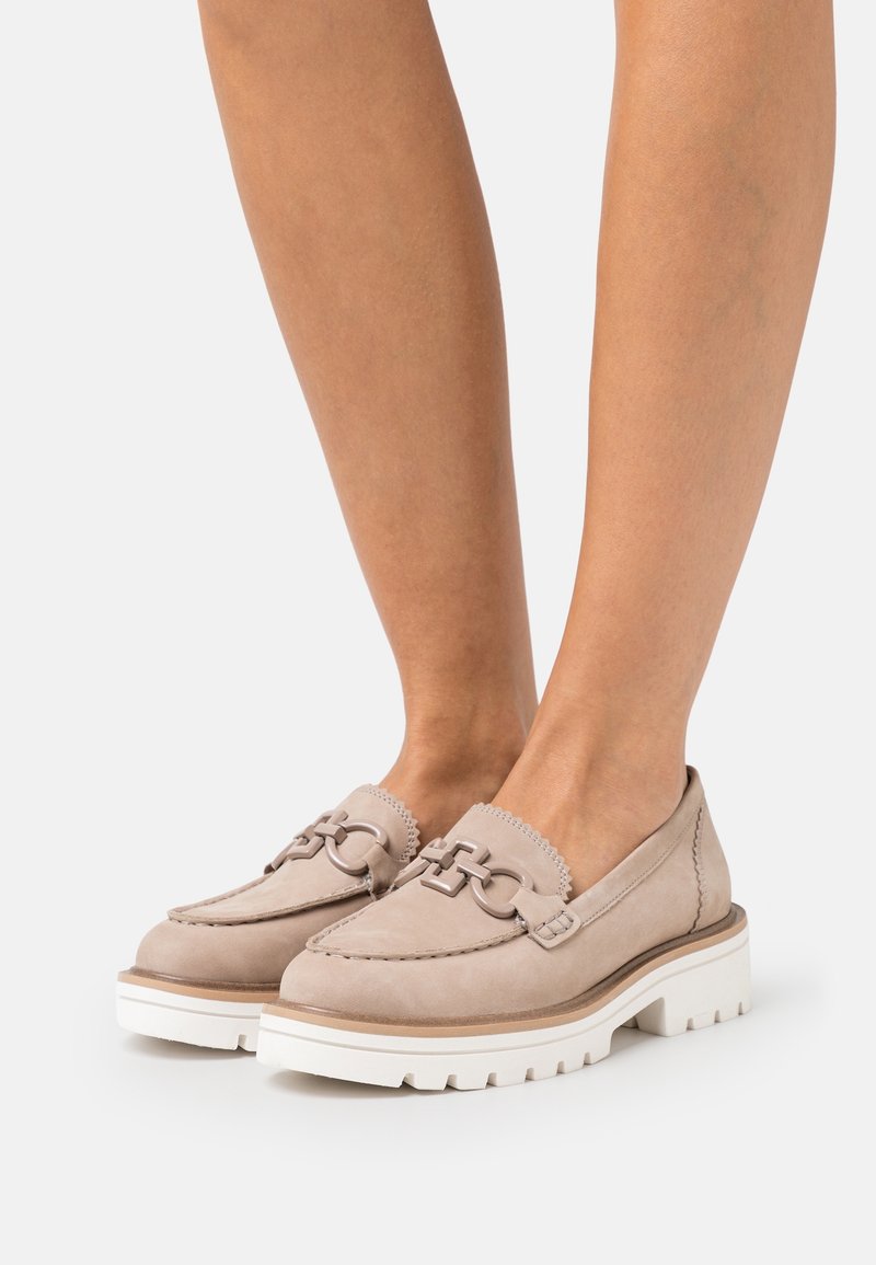 Mocassins en cuir beige avec semelles blanches épaisses et détail décoratif en chaîne métallique ronde sur des pieds aux jambes nues.