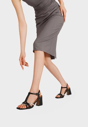 Donna che indossa un vestito grigio aderente e sandali neri con tacco alto e cinturini, in posa con un braccio teso verso il basso.