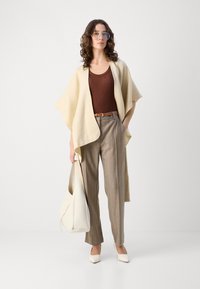 Beige lagerad sjal i kombination med en brun ribbad topp, randiga beige byxor och vita spetsiga klackskor. Håller en stor vit handväska.