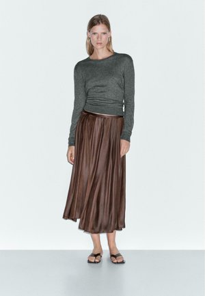 Woman stands against white background wearing gray long-sleeve top, brown pleated satin midi skirt, black sandals, and silver hoop earrings.