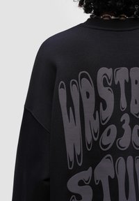 Felpa nera con vestibilità oversize, caratterizzata da una grande stampa di testo grigio distorta sul retro. Texture liscia con scollo rotondo.