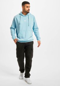 Lyseblå hættejakke med snore, frontlomme og "DEF" logo. Kombineret med sorte cargo bukser og hvide sneakers, hvilket skaber et afslappet outfit.