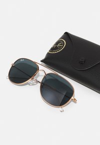 Ochelari de soare Ray-Ban cu lentile întunecate, ramă din metal auriu și suporturi nazale transparente, acompaniați de un etui texturat negru cu un logo.