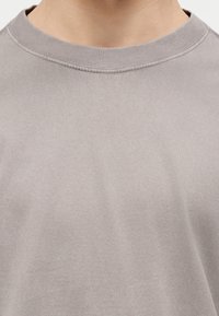 Κοντινό πλάνο ενός ατόμου που φοράει ένα απλό ανοιχτογκρίζο t-shirt με στρογγυλή λαιμόκοψη και εμφανές ράψιμο στο γιακά.