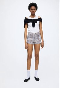 Femme aux cheveux courts et bouclés, portant un haut blanc sans manches, un short à motif paisley, un pull noir noué autour des épaules, des chaussettes blanches et des chaussures noires.