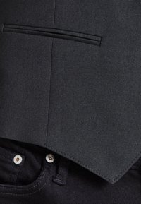 Chaleco negro texturizado con un ajuste entallado, que presenta un bolsillo estrecho, usado sobre pantalones oscuros con detalles en metal plateado.