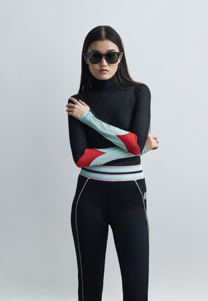 Woman with long dark hair wearing black sunglasses, black top with red and light blue geometric sleeves, and black pants with striped waistband and side seams.