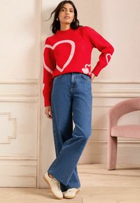 Suéter rojo con patrones de corazones rosas, combinado con jeans de pierna ancha azules y zapatillas beige. Textura suave y corte relajado.