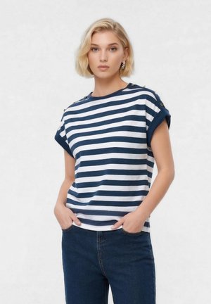 Blonde vrouw met kort haar, gekleed in een marineblauw-wit gestreept shirt met korte mouwen en donkerblauwe spijkerbroek, handen in de zakken, neutrale uitdrukking.