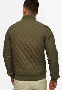 Veste bombardier matelassée vert olive avec un motif en diamant texturé, des poignets et un col côtelés, présentant une coupe ajustée et une ceinture contrastante.