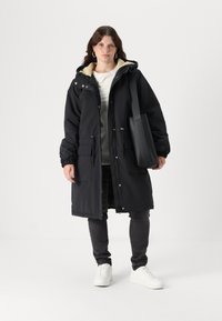Parka noire matelassée avec une capuche en fourrure, fermeture éclair et boutons pression, poches avant, associée à un jean foncé et des baskets blanches.